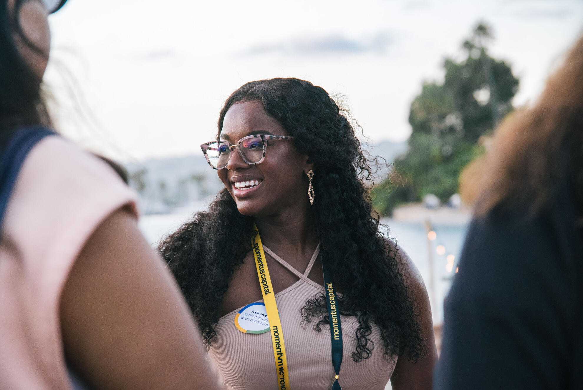 Smiling woman wearing a name badge and yellow lanyard at an outdoor professional gathering—captured candidly in golden-hour light with authentic energy and warmth.